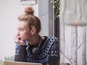 Pensive young woman with headphones at laptop looking out cafe window