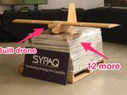 組み立てられたSYPAQのドローンが、平らに梱包された状態のドローンの積み重ねの上に置かれている。