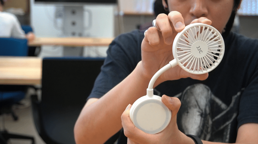 A person is holding a small, portable white fan with a flexible neck and circular blades in front of them. The fan has the letters 