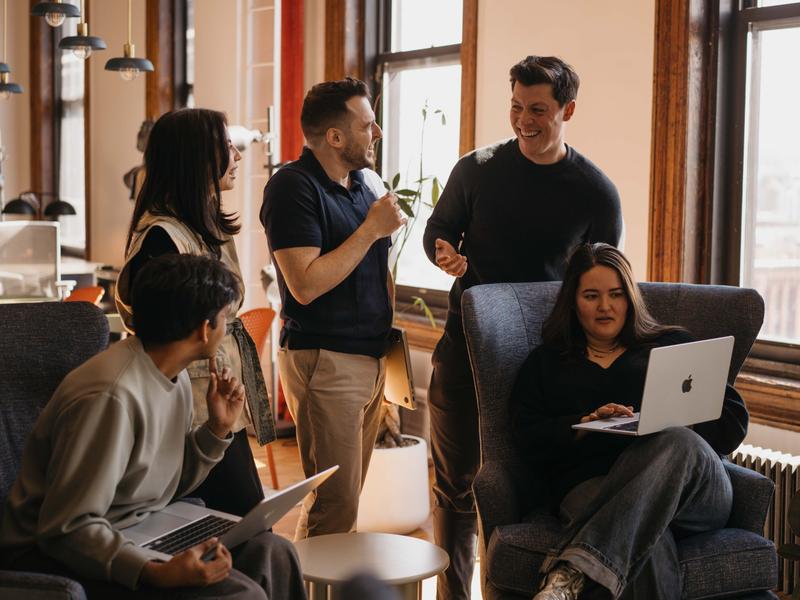 Coworkers chat and smile in a sunlit office lounge while two people use laptops in armchairs by the windows.