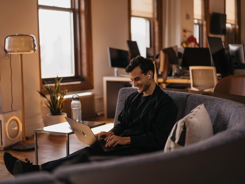 A man wearing earbuds smiles while working on a laptop from a couch in a quiet, sunlit office.