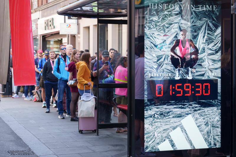 People queueing by a bus stop which displays an Adidas ad of Sabastian Sawe and his record marathon time.