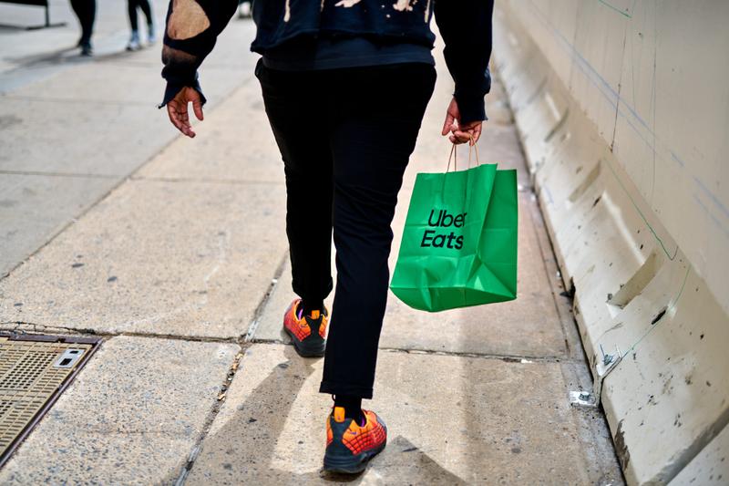 A person walks away from the camera carrying a green paper Uber Eats bag while wearing dark pants and red, yellow, and black sneakers.