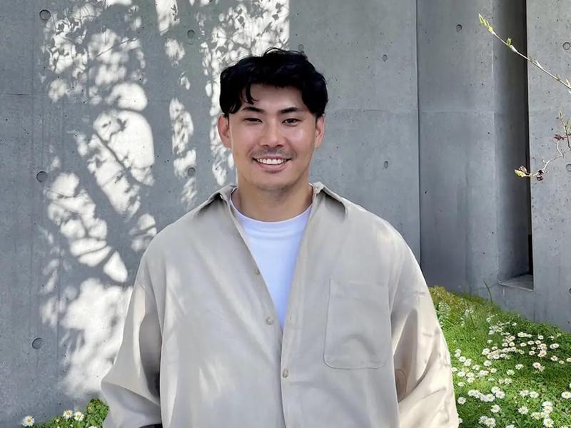 A man wearing a beige shirt standing in front of a concrete wall.