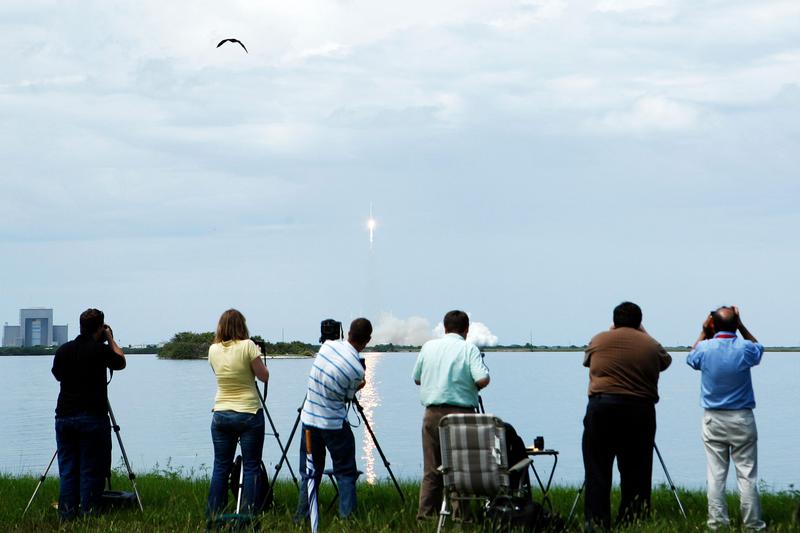 Onlookers take pictures from across a waterway while a SpaceX Falcon 9 takes off.