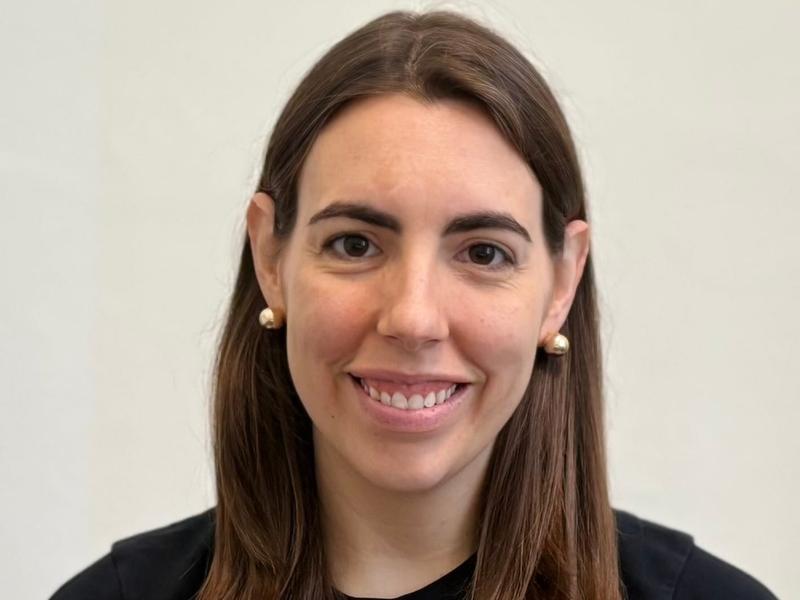 Smiling person with shoulder-length brown hair wearing a black top against a plain light background.