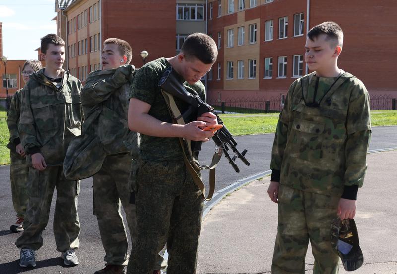 A Russian teenager looks at his phone while holding a Kalashnikov rifle.